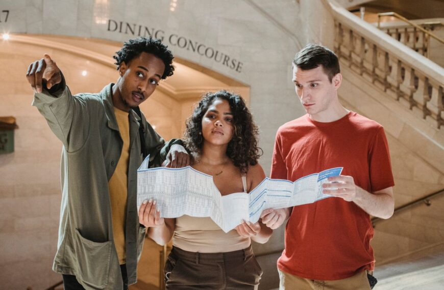 Young African American male tourist pointing away while searching for direction with diverse fiends standing in railway station terminal with paper map in hands