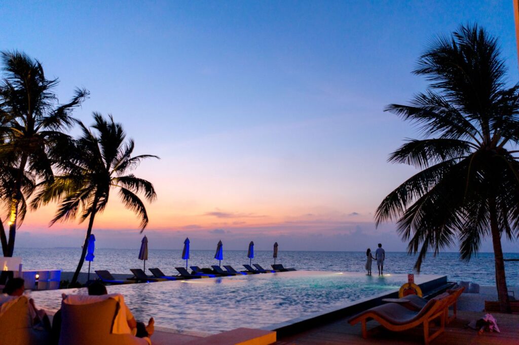 Et par nyder en stemningsfuld solnedgang ved en infinity-pool ved havet omgivet af palmer.