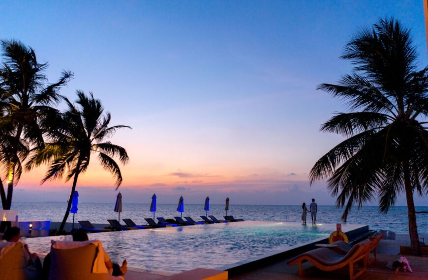 A couple enjoys a picturesque sunset by the seaside infinity pool, surrounded by palm trees.