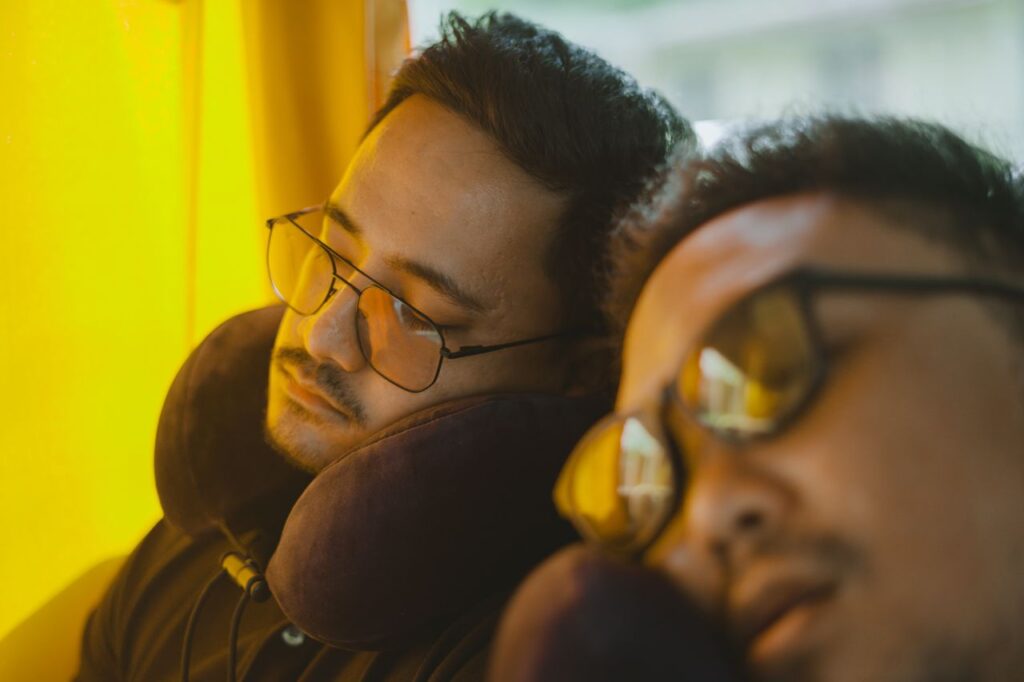 Mænd med briller sover i en bus med nakkepuder for ekstra komfort under rejsen.

