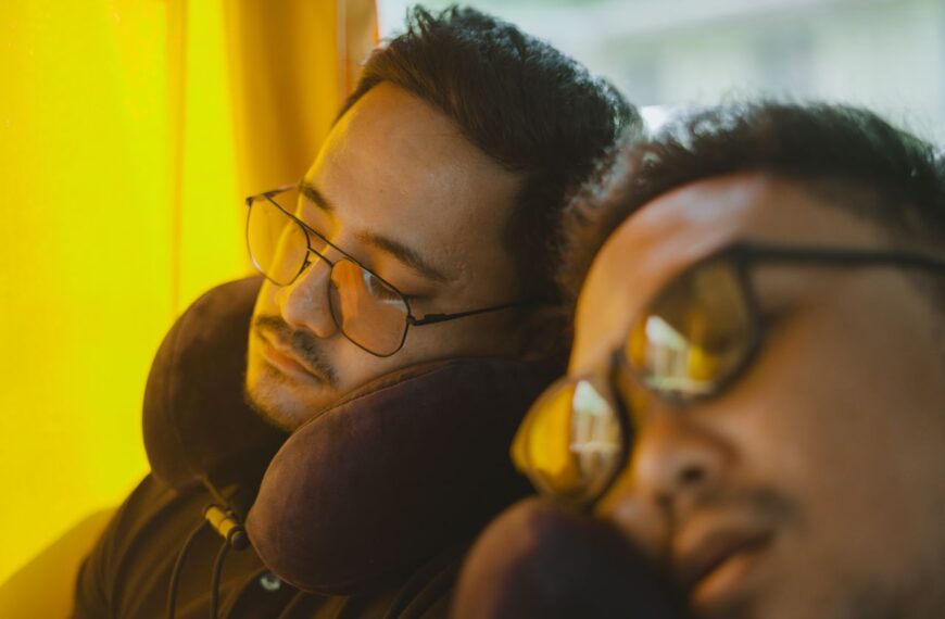 Two men wearing eyeglasses sleep on a bus, using neck pillows for comfort during the journey.