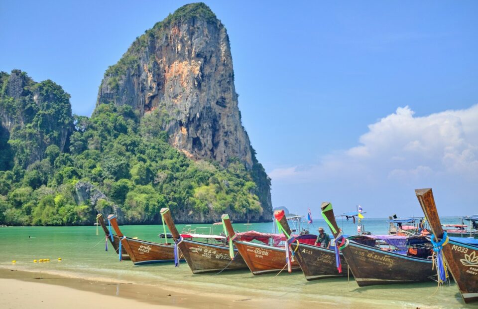 Langhalebåde ved en sandstrand foran grønklædte kalkstensklipper i Krabi