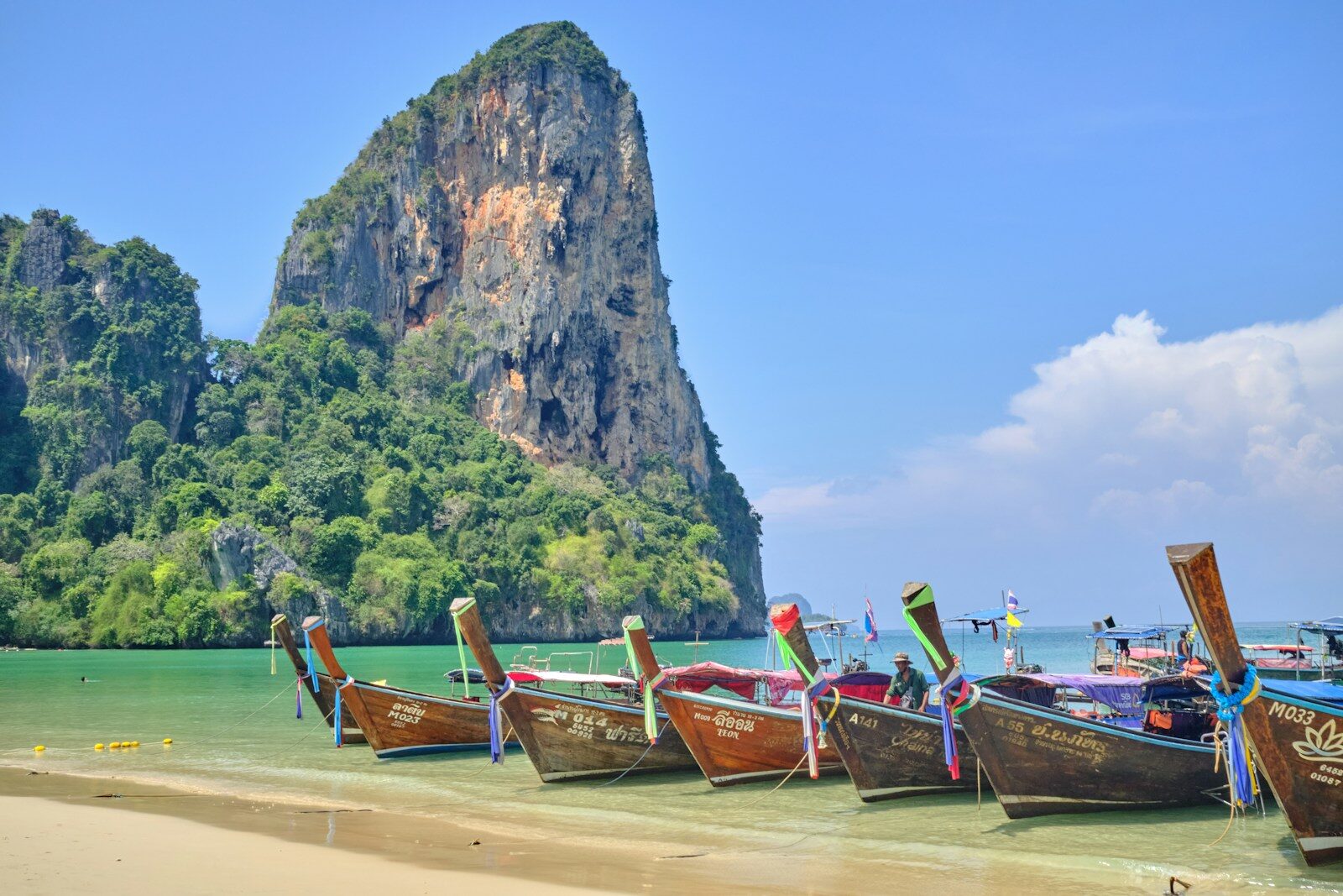 Langhalebåde ved en sandstrand foran grønklædte kalkstensklipper i Krabi